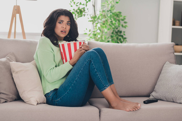 Full body profile side photo of dark skin young woman sit couch scared watch horror film eat pop corn indoors in house - Foto, Imagem