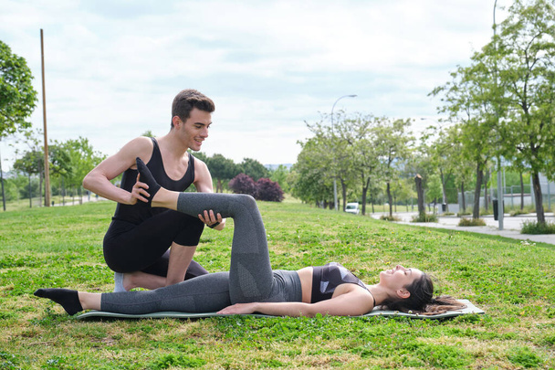 Personal trainer helping young woman in a park with stretching exercises. Leg stretch laying on a mat outside. - Foto, Imagem