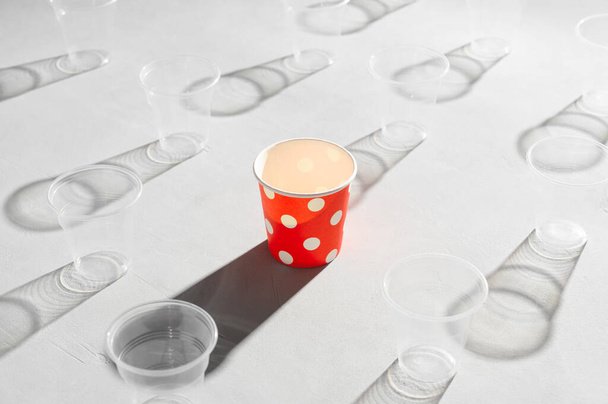Környezetbarát piros papírpohár körül műanyag poharak egy szürke asztalon. Műanyagmentes fogalom, biológiailag lebomló asztali áruk, újrahasznosítható. - Fotó, kép