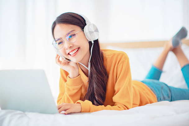 Mulher feliz relaxando ao usar fones de ouvido assistindo vídeos no laptop deitado em uma cama em casa, Relaxe e conceito de lazer. - Foto, Imagem