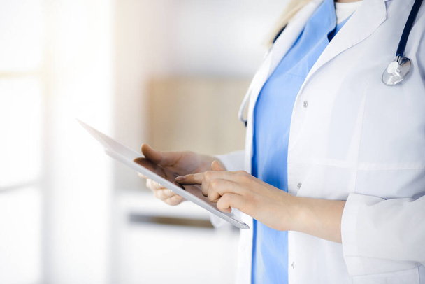 Unknown woman-doctor standing in sunny clinic and using tablet pc, close-up. Data and best service in medicine - Foto, Imagen