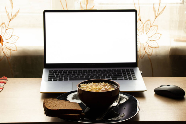 a plate of noodles stands next to a laptop with a white screen on the table in the room, the windows were taken, lunch at the desk at home, food - Photo, image