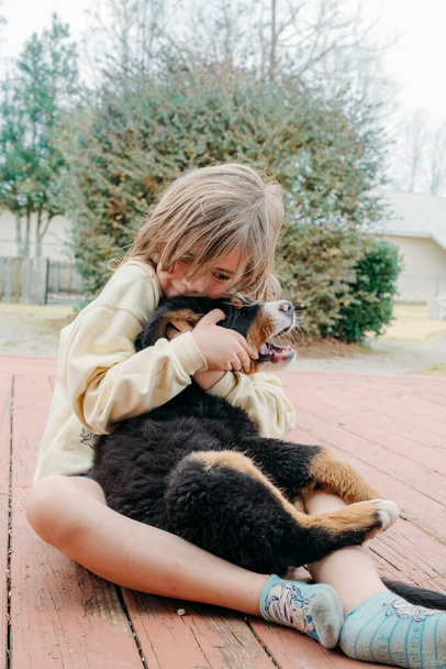 Αξιολάτρευτο χαριτωμένο κοριτσάκι κάθεται με οικόσιτα σκυλιά και αγκαλιάζει μικρό κουτάβι Bernese στη βεράντα του σπιτιού. Σκύλοι. Έννοια της αγάπης για τα ζώα και φιλία με τα κατοικίδια ζώα. - Φωτογραφία, εικόνα