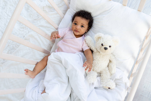 boldog mosolygós afro-amerikai baba egy kiságyban egy plüssmackóval elalszik, vagy megy aludni - Fotó, kép
