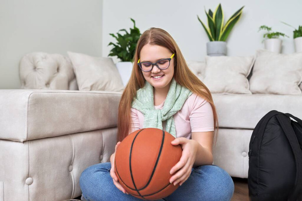 Happy smiling teenage girl with basketball ball in hands sitting on floor at home - Фото, зображення