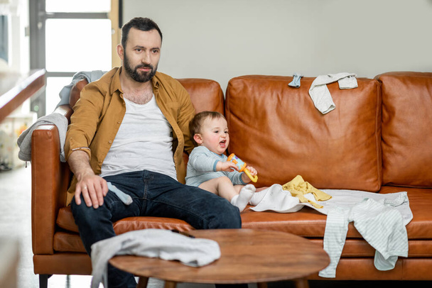 Zmęczony ojciec karmiący noworodka siedząc na kanapie - Zdjęcie, obraz