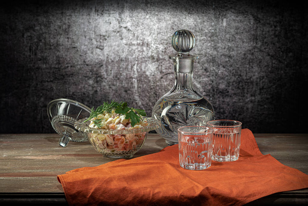 Salad bowl with olivier salad, with glass and decanter, red napkin, on a wooden table top, on a gray background - Fotoğraf, Görsel