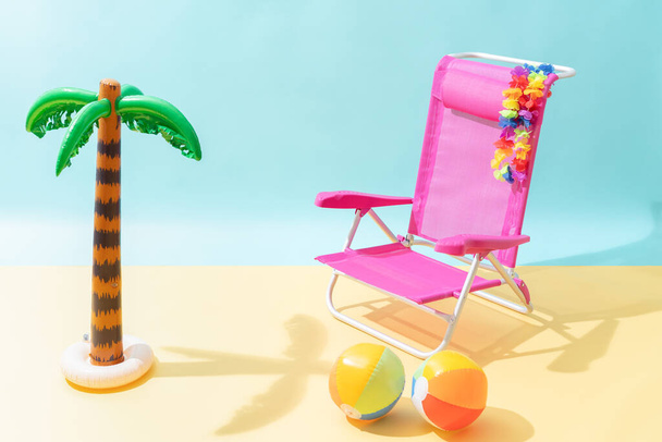 Egy rózsaszín tengerparti szék két színes strandlabdával, kék és sárga háttérrel. A székre akasztanak egy virágos nyakláncot. A szék egy felfújható pálmafa árnyékában van.. - Fotó, kép