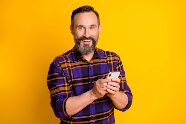 Photo portrait of happy smiling man keeping mobile phone reading news isolated on vivid yellow color background - Foto, Imagem