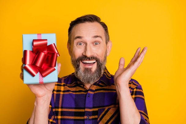 Photo portrait of happy smiling man showing wrapped present on birthday party surprised isolated on vibrant yellow color background - Photo, image