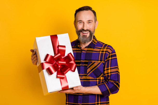 Portrait of attractive cheerful guy wearing checked shirt holding in hands bow ribbon box isolated over bright yellow color background - Fotó, kép