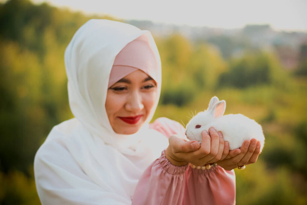 Muslim woman hold a small white rabbit in her arms. A portrait of a Muslim woman in a pink dress and a scarf, a pearl bracelet. Tenderness. - Foto, imagen