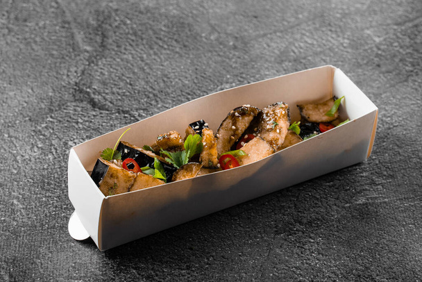 Fried eggplant dish in cardboard box. Food delivery. Vegetables garnished with cilantro and chili on gray background - Photo, Image
