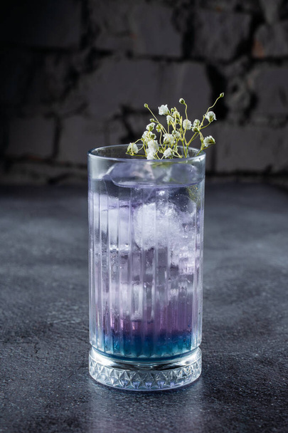 Egy koktél egy magas pohárban, kék likőrrel és jéggel, fehér virágokkal díszítve, szürke alapon. - Fotó, kép