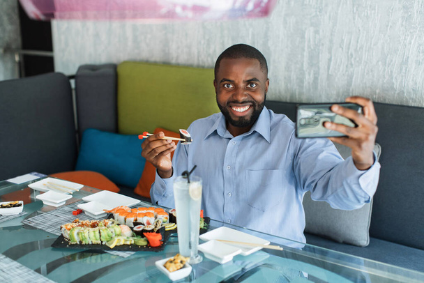 Задоволений усмішкою молодого африканського бізнесмена, який робить самовідтворення або відеодзвінок на мобільний телефон, тримаючи палички суші рулон, традиційний східний обід в ресторані.. - Фото, зображення