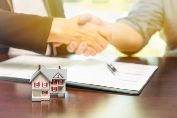 Agent nieruchomości i klient podpisują umowy i uścisk dłoni z małym modelem domu z przodu. - Zdjęcie, obraz