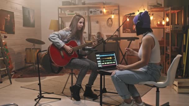 Полноценное медлительное движение девушек в рок-одежде, создающих новую сессию записи песен на акустической гитаре, сидящих в уютной музыкальной студии старой школы с ноутбуком - Кадры, видео
