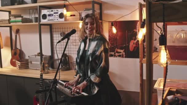 Medium slowmo van koele energieke jonge vrouw in geblokte shot spelen elektronische piano en zingen in microfoon in gezellige retro-stijl studio - Video