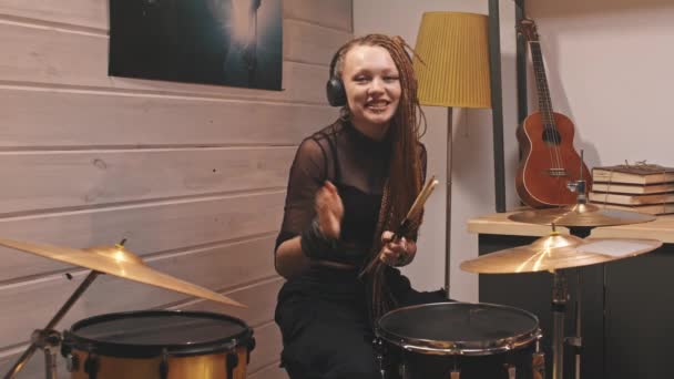 Medium shot of young woman in rock-style clothes with bright makeup and dreadlocks saying hello to camera, putting on headphones and starting playing drums in cozy music studio - 映像、動画