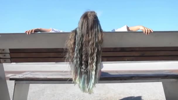 Brünettes preteen Mädchen mit blauen Farbakzenten sitzt auf Bank auf Brücke, Rückansicht, Sommertag - Filmmaterial, Video
