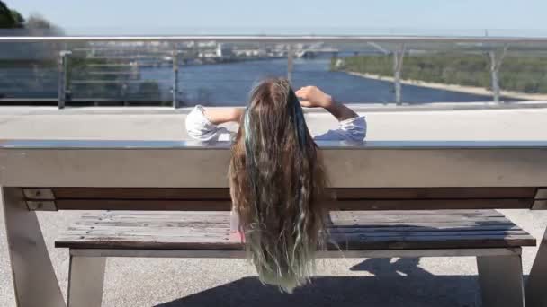 Morena chica preadolescente con reflejos de color azul sentado en el banco en el puente, vista trasera, día de verano - Imágenes, Vídeo