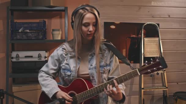 Medium shot of young smiling woman in vintage denim jacket saying hello to camera, putting on headphones and starting playing acoustic guitar in cozy retro-style music studio - 映像、動画