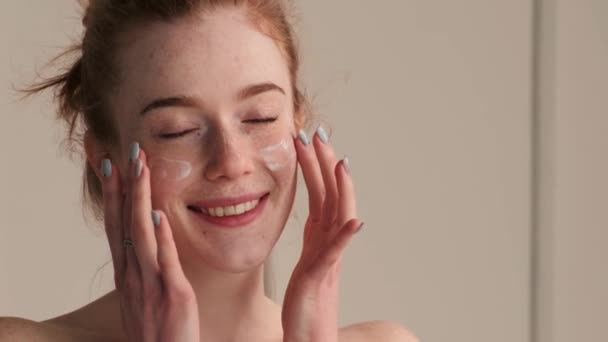 donna con i capelli rossi si sta massaggiando il viso con crema anti invecchiamento sorridente alla fotocamera - Filmati, video