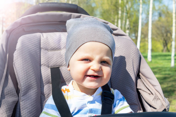 Adorable happy caucasian baby boy sitting in grey stroller in park. Summer day. Child is strapped in stroller during the walk.  - Foto, imagen