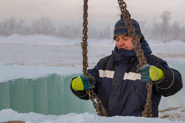 Portret van een arbeidersslinger in een blauw jasje die ijsblokken uitlaadt met behulp van een kettingspin - Foto, afbeelding