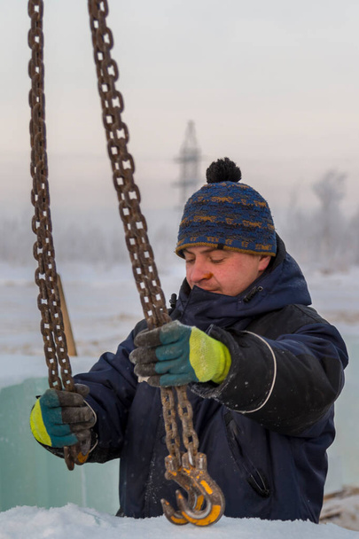Retrato de un tirador trabajador en una chaqueta azul descargando bloques de hielo usando una araña de cadena - Foto, Imagen