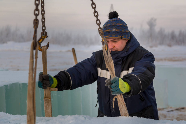 Portrait d'un travailleur fronçant dans une veste bleue déchargeant des blocs de glace à l'aide d'une araignée à chaîne - Photo, image