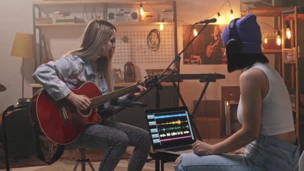 Střední zpomalení mladé ženy hudebnice hraje na akustickou kytaru, zatímco její kolegyně ve sluchátkách pomocí notebooku pro nahrávání relace v retro stylu hudebního studia - Záběry, video
