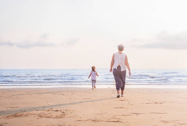 Babička a dítě jdou při západu slunce po pláži. Dívka utíká od ženy k vodě - Fotografie, Obrázek