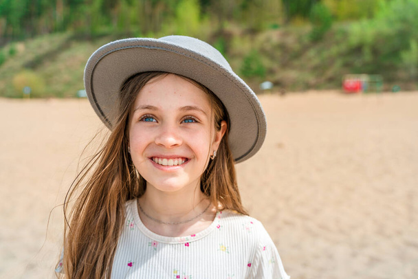 Portrait of a smiling little girl in a wide-brimmed hat standing on a sandy beach by the sea - Photo, Image