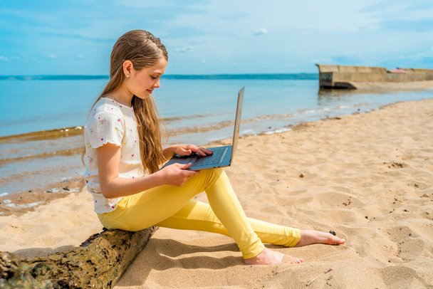 Ein kleines Mädchen mit langen Haaren sitzt auf einem Baumstamm am Strand, einen Laptop auf dem Schoß. Das Kind benutzt einen Laptop - Foto, Bild