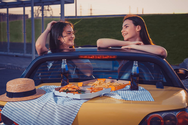 Mooie modellen glimlachen, poseren in een gele auto met frietjes, pizza, hoed en frisdrank in glazen flessen op de kofferbak. Fast food. Sluiten, kopieerruimte - Foto, afbeelding