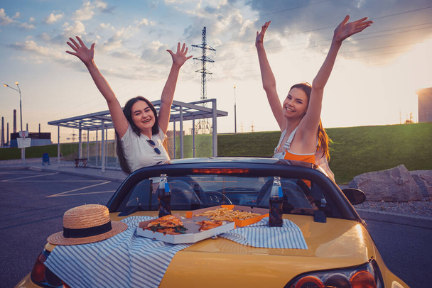 Дамы в повседневной одежде улыбаются, поднимают руки, позируют в жёлтой машине с картошкой фри, пиццей и содовой на багажнике. Фастфуд. Копирование пространства - Фото, изображение
