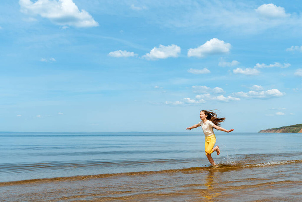 Маленька дівчинка в яскраво-жовтих штанях біжить у воді і фролі на пляжі
 - Фото, зображення