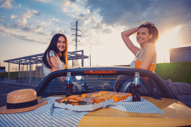 Amigos hembras sonríen, posando en coche amarillo cabrio con papas fritas, pizza y refrescos en botellas de vidrio en su maletero. Comida rápida. Copiar espacio - Foto, Imagen