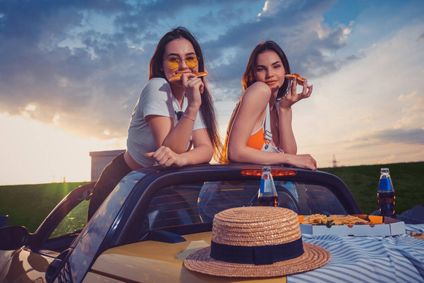 Charmante meisjes die pizza eten, poseren in een gele auto roadster met frietjes, hoed en frisdrank in glazen flessen op de kofferbak. Fast food. Zwijg. - Foto, afbeelding