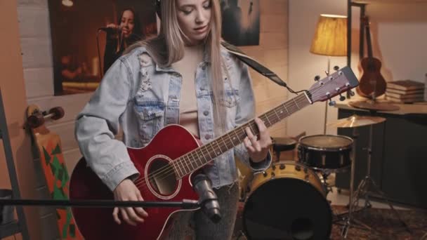 Портрет молодої усміхненої жінки-гітариста з довгим світлим волоссям у старовинній джинсовій куртці, що дивиться на камеру, граючи на акустичній гітарі в затишній музичній студії в стилі ретро
 - Кадри, відео