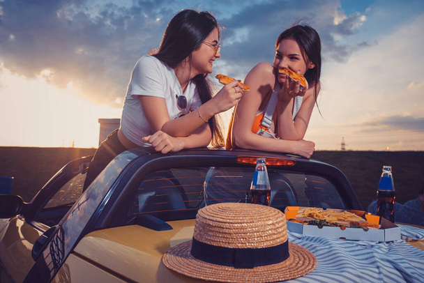 Duas jovens estão comendo pizza, rindo, posando em carro amarelo com batatas fritas, chapéu e refrigerante em garrafas de vidro no tronco. Comida rápida. Faz asneira. - Foto, Imagem