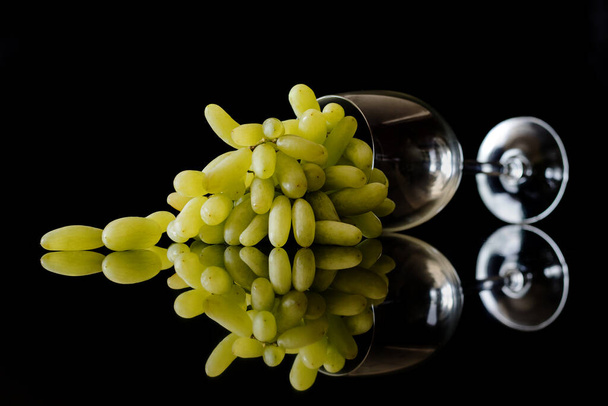racimo de uvas blancas en una clase de vino sobre una superficie negra reflejada  - Foto, Imagen
