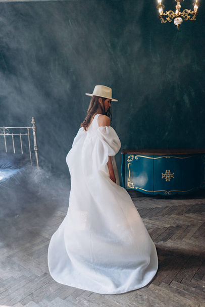 girl in a beautiful white sensual dress and white hat standing in the blue room and turning with her back to the camera - Foto, Imagem