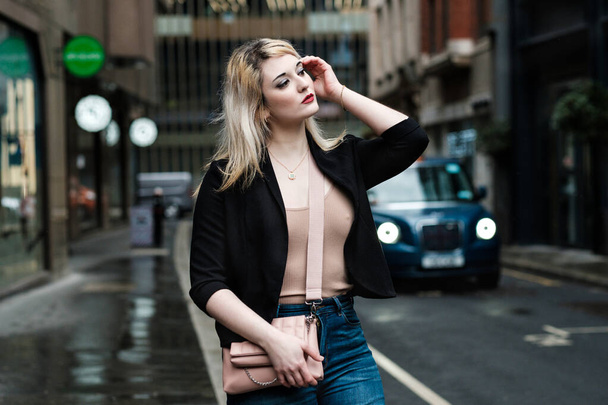 Urban girl in London, she wears black jacket, jeans, cream t-shirt and bag. Cloudy day. Man and taxi on the background. Taxi behind. - Foto, imagen