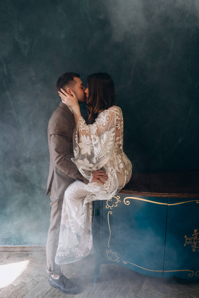 joven novio abrazando y besando a su hermosa novia vestida con un vestido blanco bohemio en una habitación humeante - Foto, Imagen