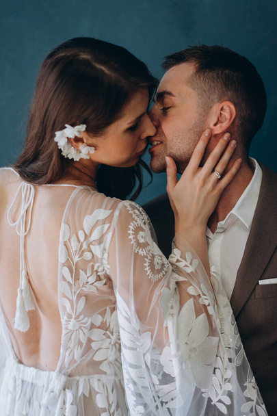 the beautiful young bride dressed in a white bohemian dress standing in half-turn and kissing her groom dressed in the elegant suit in the blue room - Фото, изображение