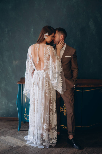 the beautiful young bride dressed in a white bohemian dress standing in half-turn and kissing her groom dressed in the elegant suit in the blue room - Foto, Imagen