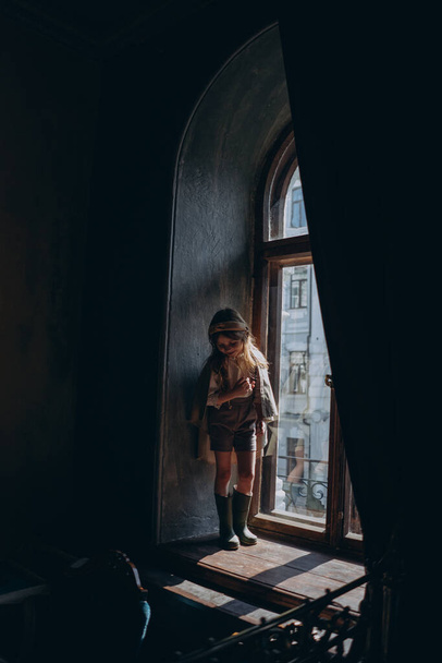 the child in vintage clothes, the little girl in a beige riding hood, shorts, and green rubber boots standing with her head down at the big arch window with curtains - Φωτογραφία, εικόνα