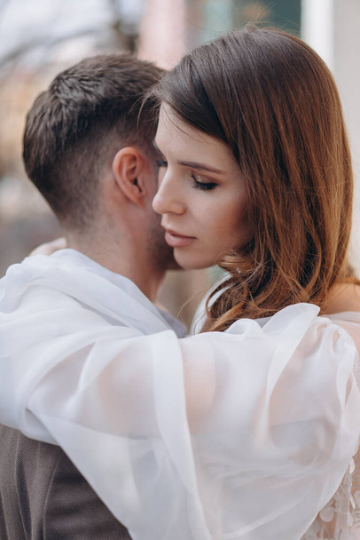 girl dressed in a beautiful white sensual tulle dress standing on the balcony and hugging her man dressed in the elegant suit, closeup view - Foto, Bild